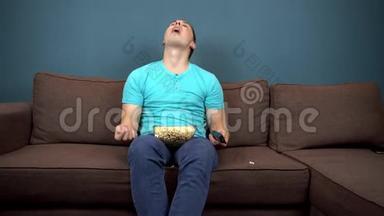 一个年轻人正在看电视和吃爆米花。 那家伙用嘴抓爆米花。 坐在沙发上。 观点